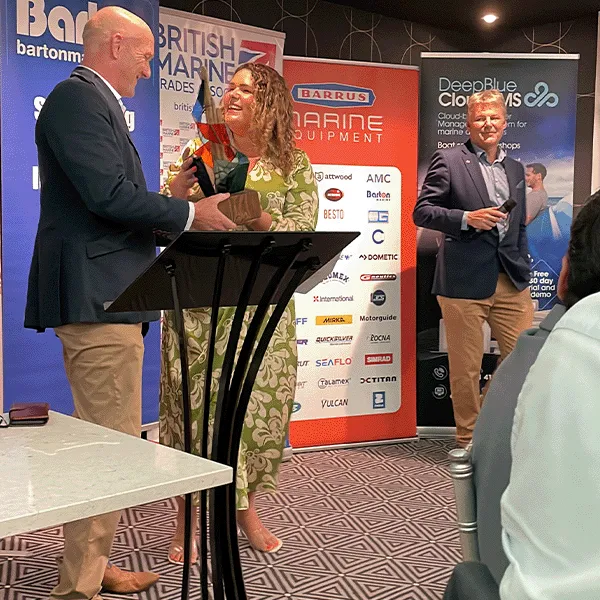 A woman in a green dress receives a trophy from a bald man while another man stands beside a banner in the background.