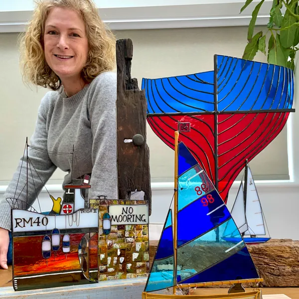 A person smiling behind colorful nautical-themed stained glass artworks, including boats and a building with the sign 