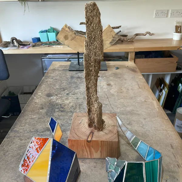 A sculpted wooden piece stands upright on a wooden base, surrounded by colorful geometric stained glass pieces on a table.