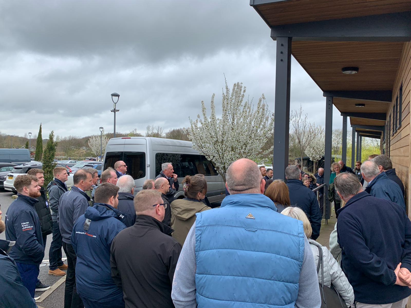 A group of people gathered outside a building, standing near a van in a parking lot under a cloudy sky.