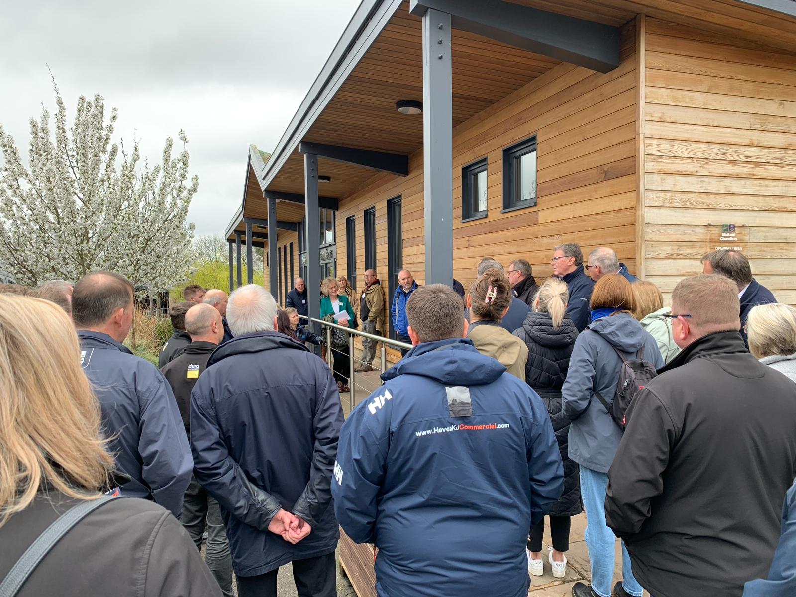 A group of people stands outside a wooden building, listening to someone speak on a platform on a cloudy day.