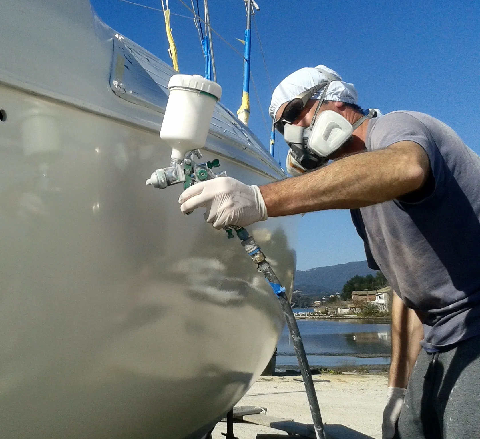 A person wearing protective gear uses a spray gun to paint a white boat on a sunny day near a body of water.