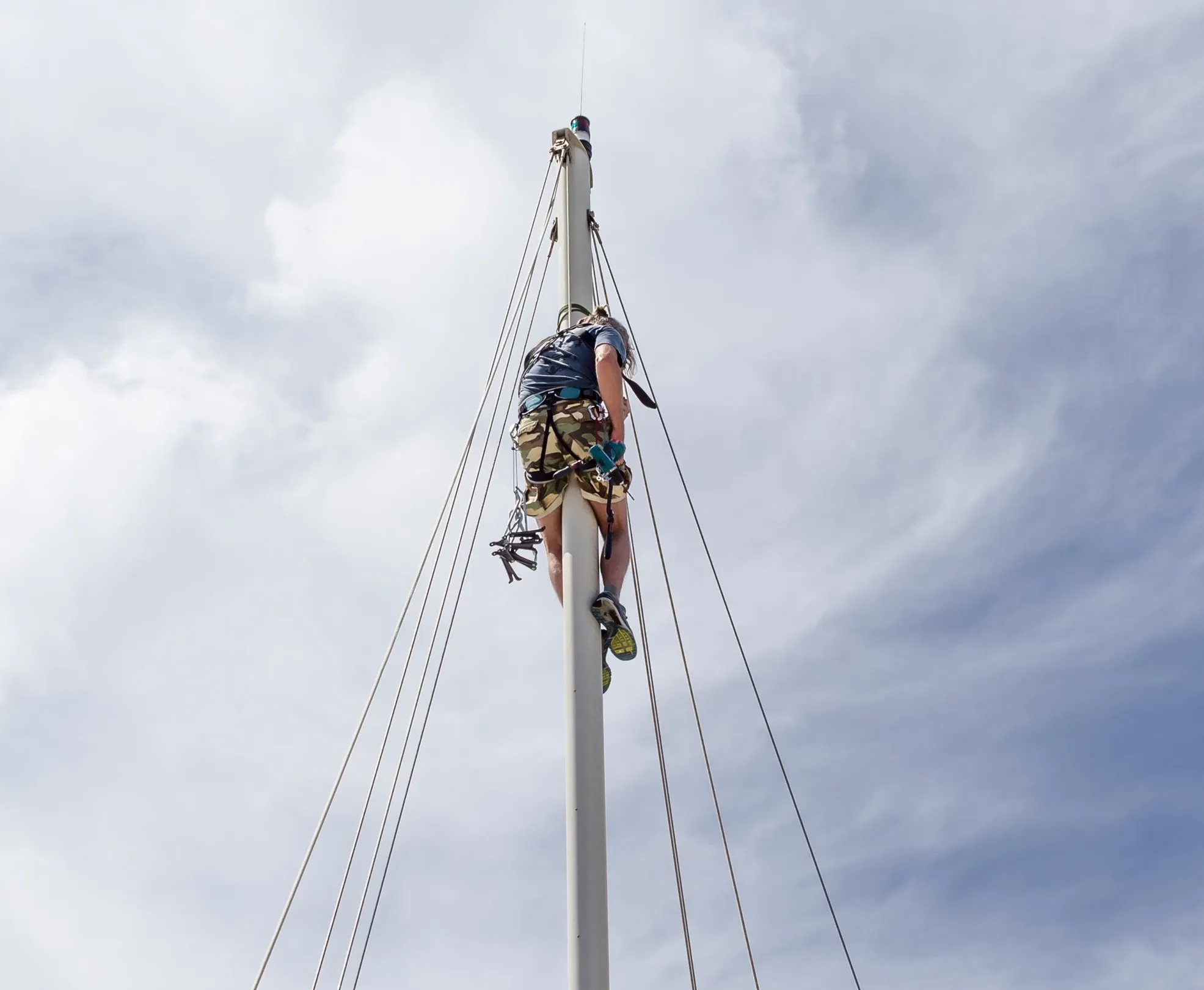 A person wearing climbing gear is ascending a tall mast against a backdrop of cloudy sky.