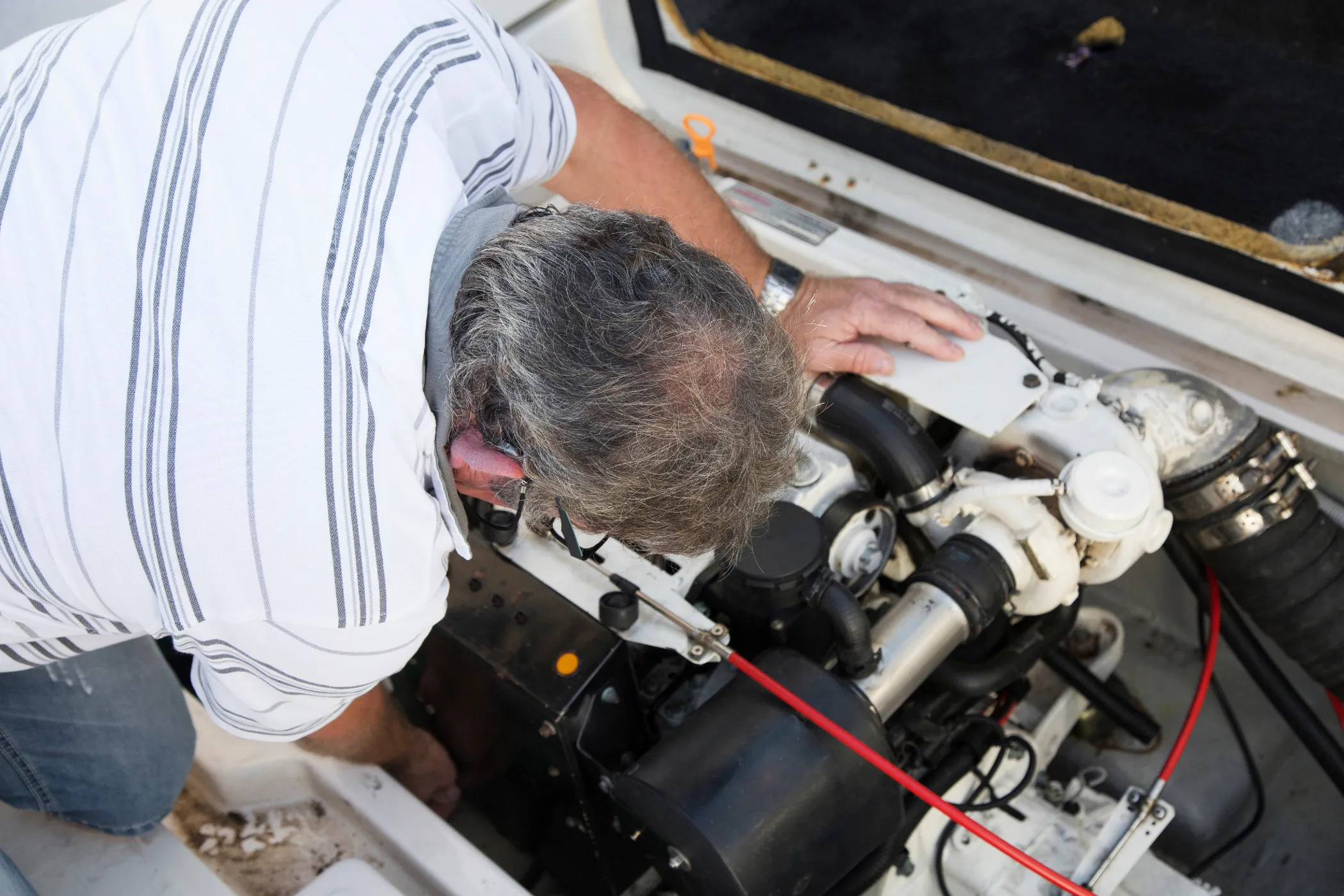 A man in a striped shirt is inspecting and working on a boat engine. He is bending over, focusing on the engine components.