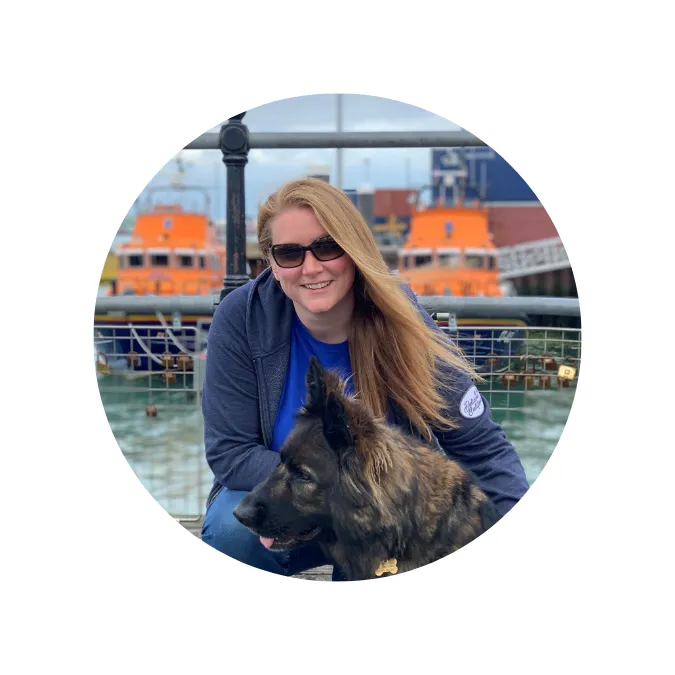 Woman with long hair wearing sunglasses and a blue jacket, kneeling next to a large dark dog near a waterfront with orange boats in the background.