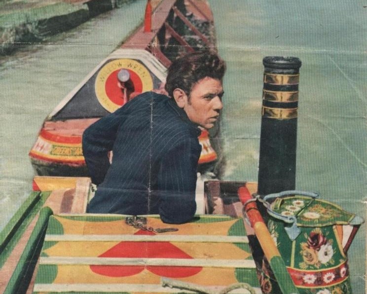 A man with dark hair navigates a narrowboat on a canal, looking towards the back. The boat's deck features colorful decorations.