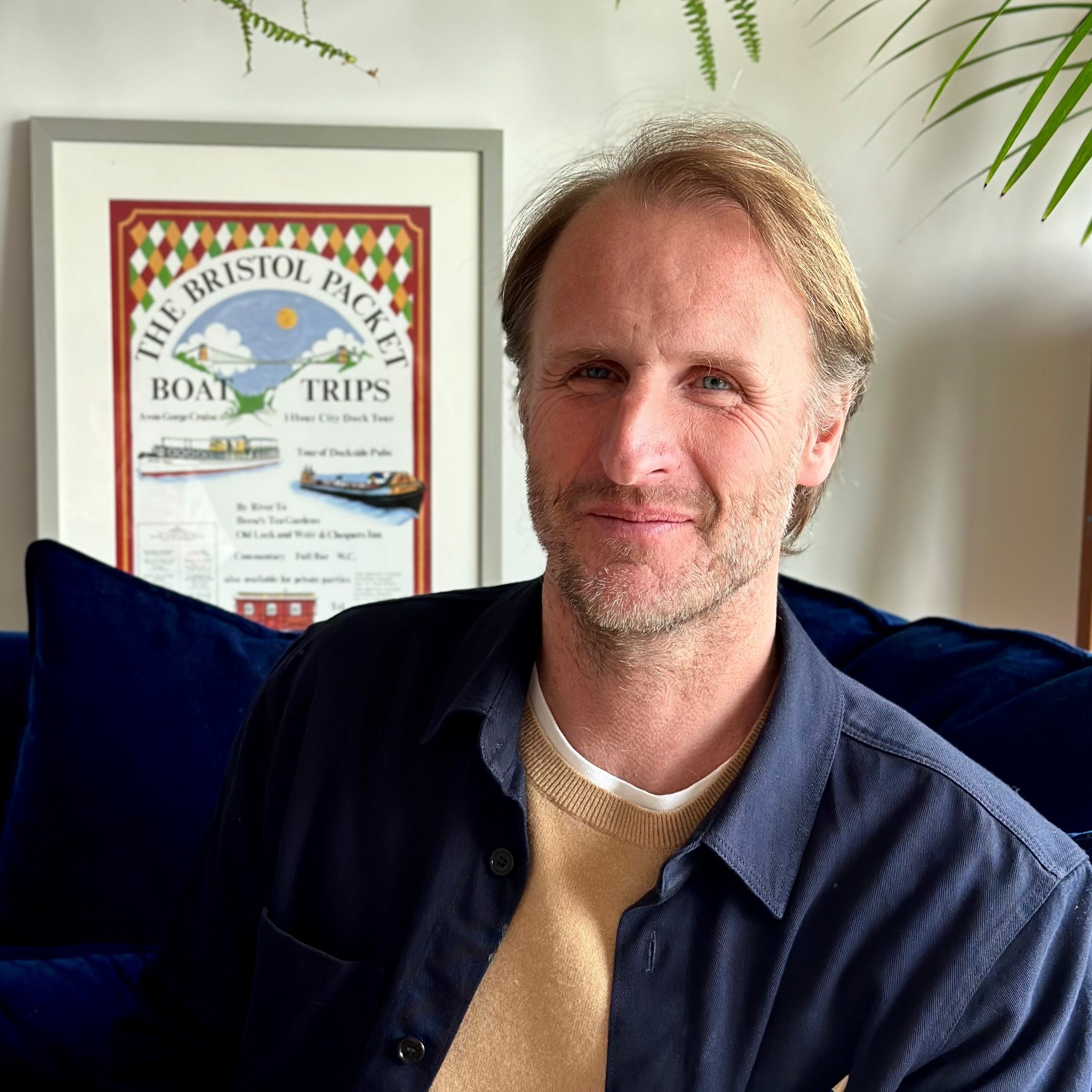 A man with light brown hair and a beard sits on a blue couch, smiling. A framed boat trips poster is on the wall behind him.