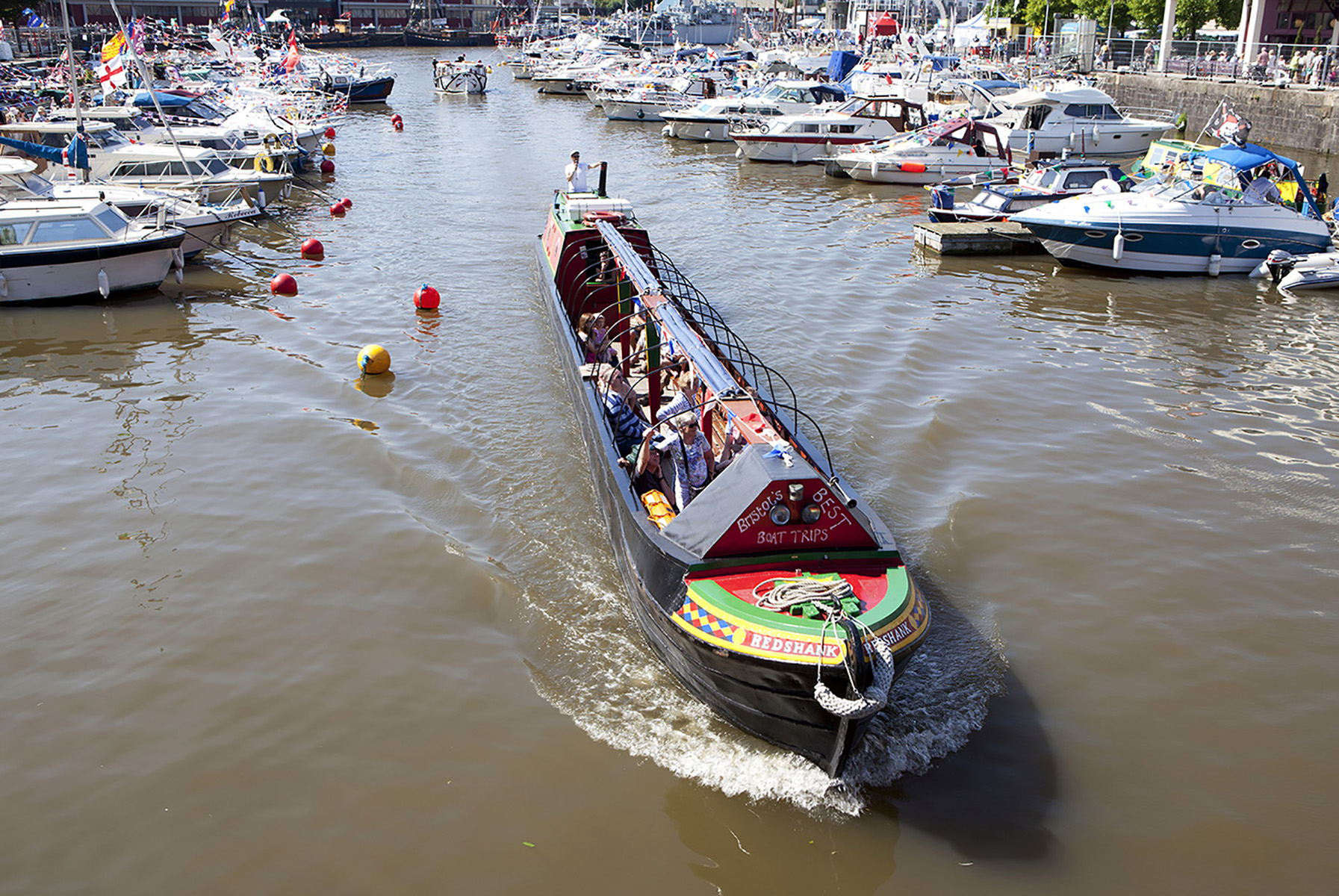 Narrow boat with people on board navigates through a marina filled with various small boats on a sunny day.