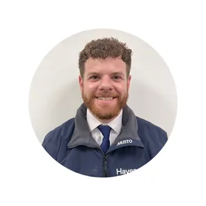 Man with curly hair and a beard, wearing a jacket and tie, smiling in front of a plain white background.